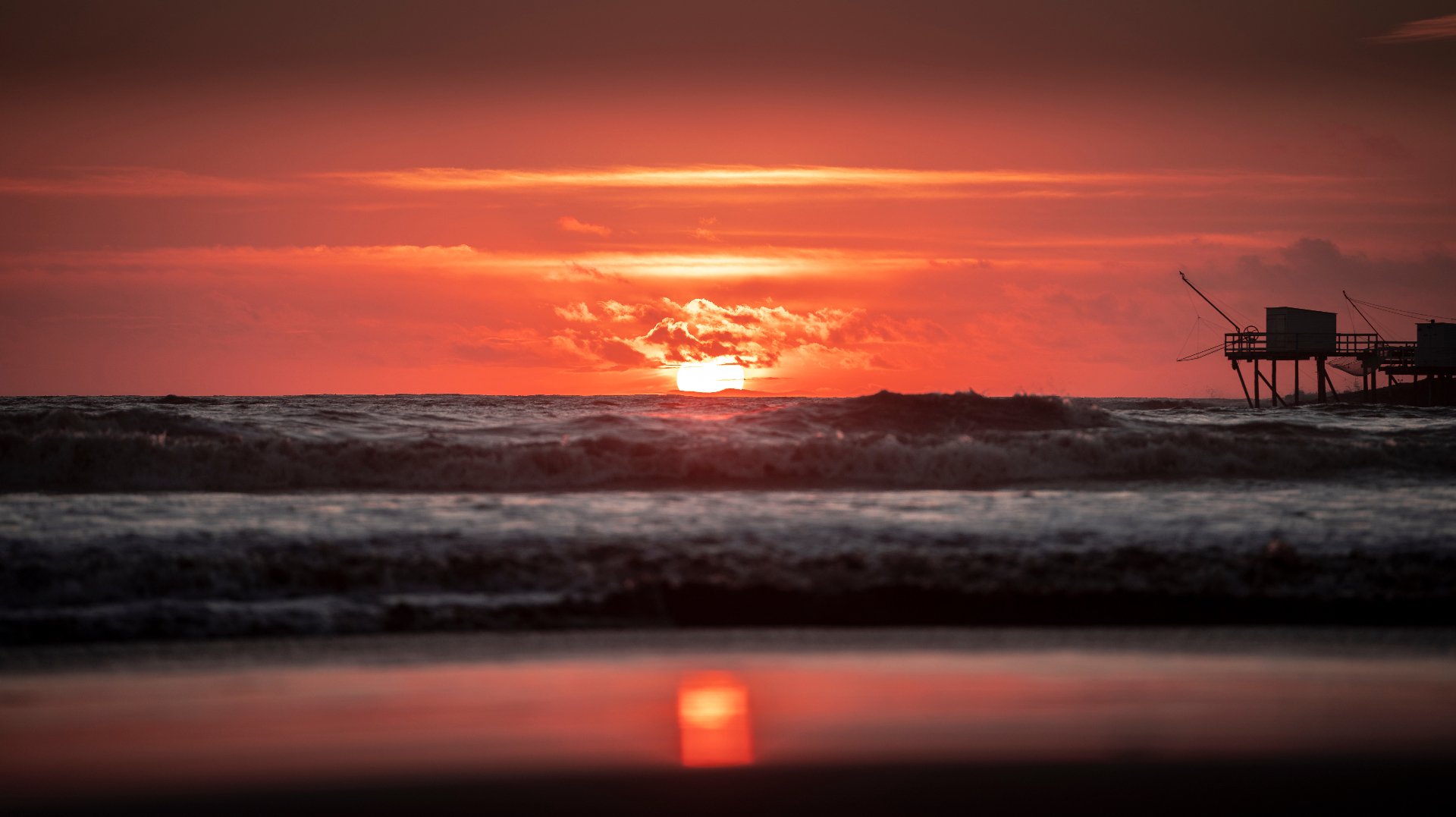 Soleil rouge couchant derrière un carrelet de Charente-Maritime, vagues au premier plan
