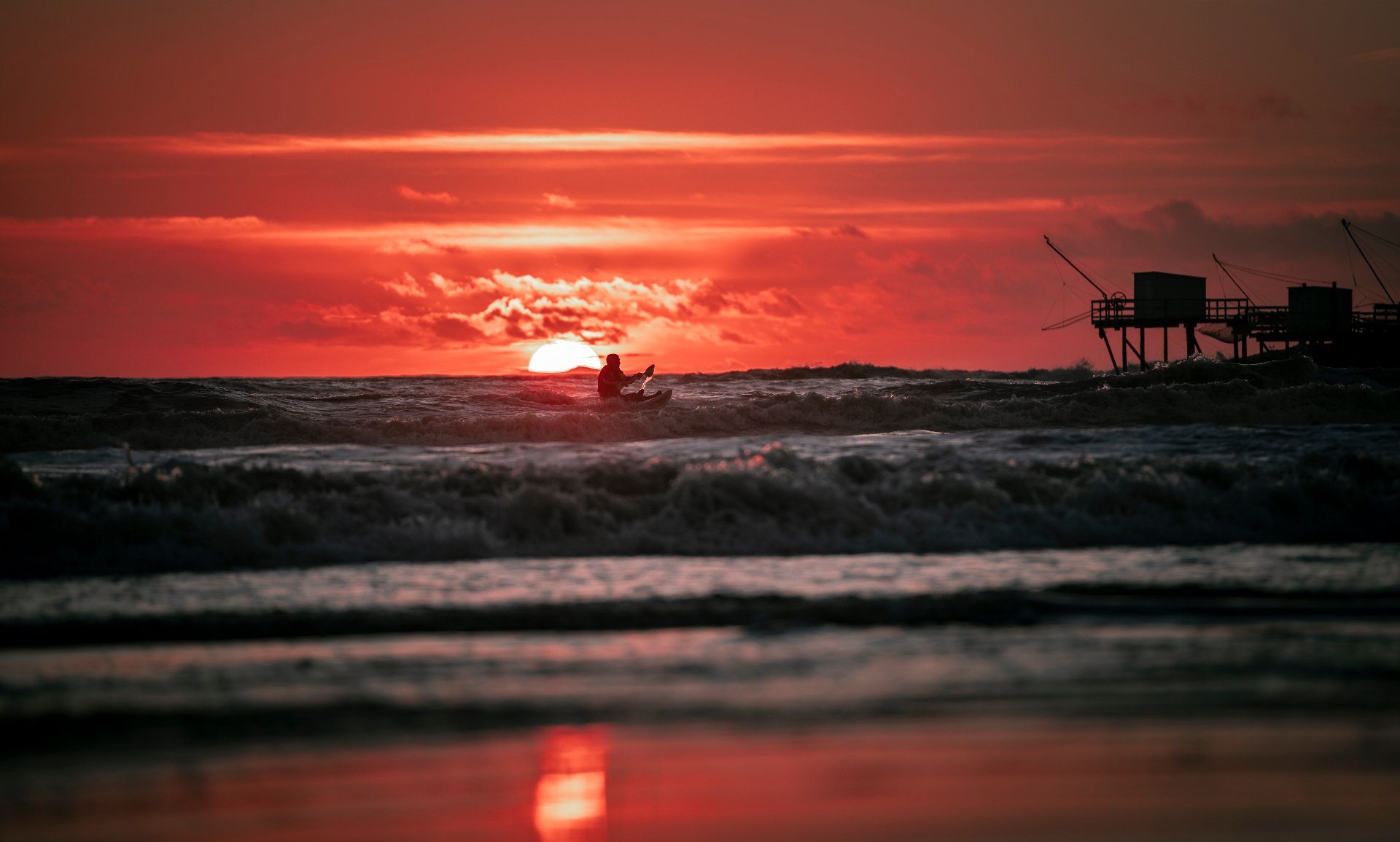 Kayakiste en silhouette sur les vagues au soleil couchant, carrelet à l'horizon